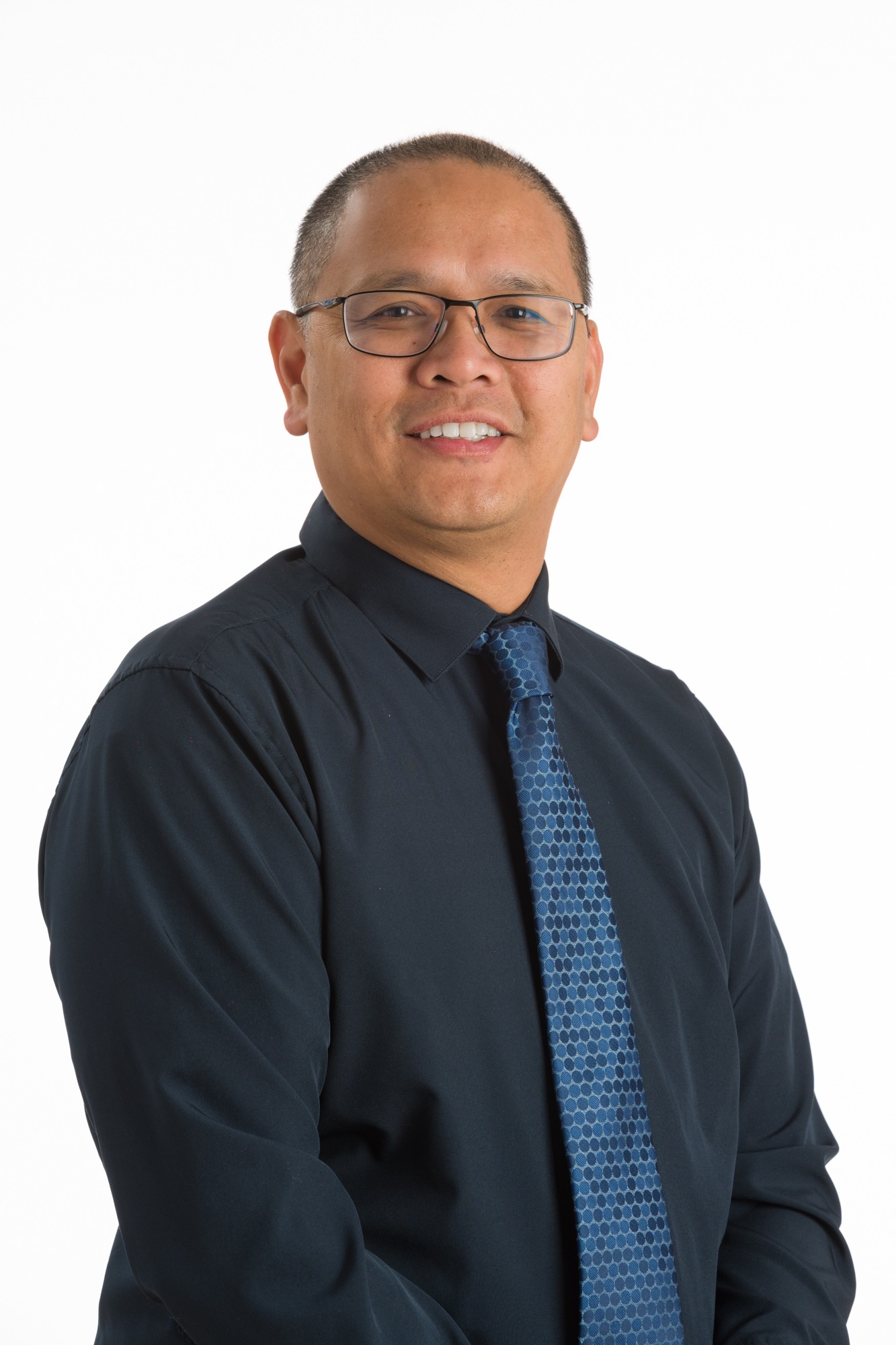 A man with short hair and glasses, wearing a dark shirt and patterned blue tie, poses against a plain white background.