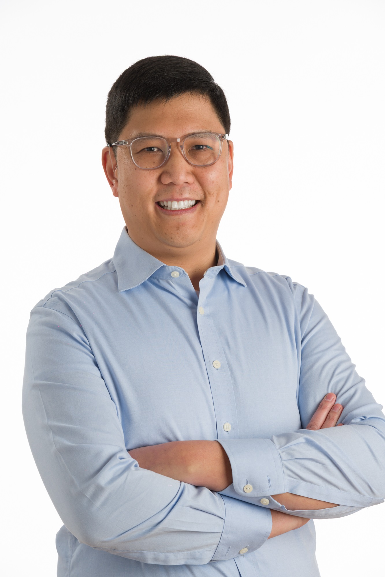 Christopher Chin, wearing glasses and a light blue button-up shirt, stands with arms crossed, smiling against a plain white background.