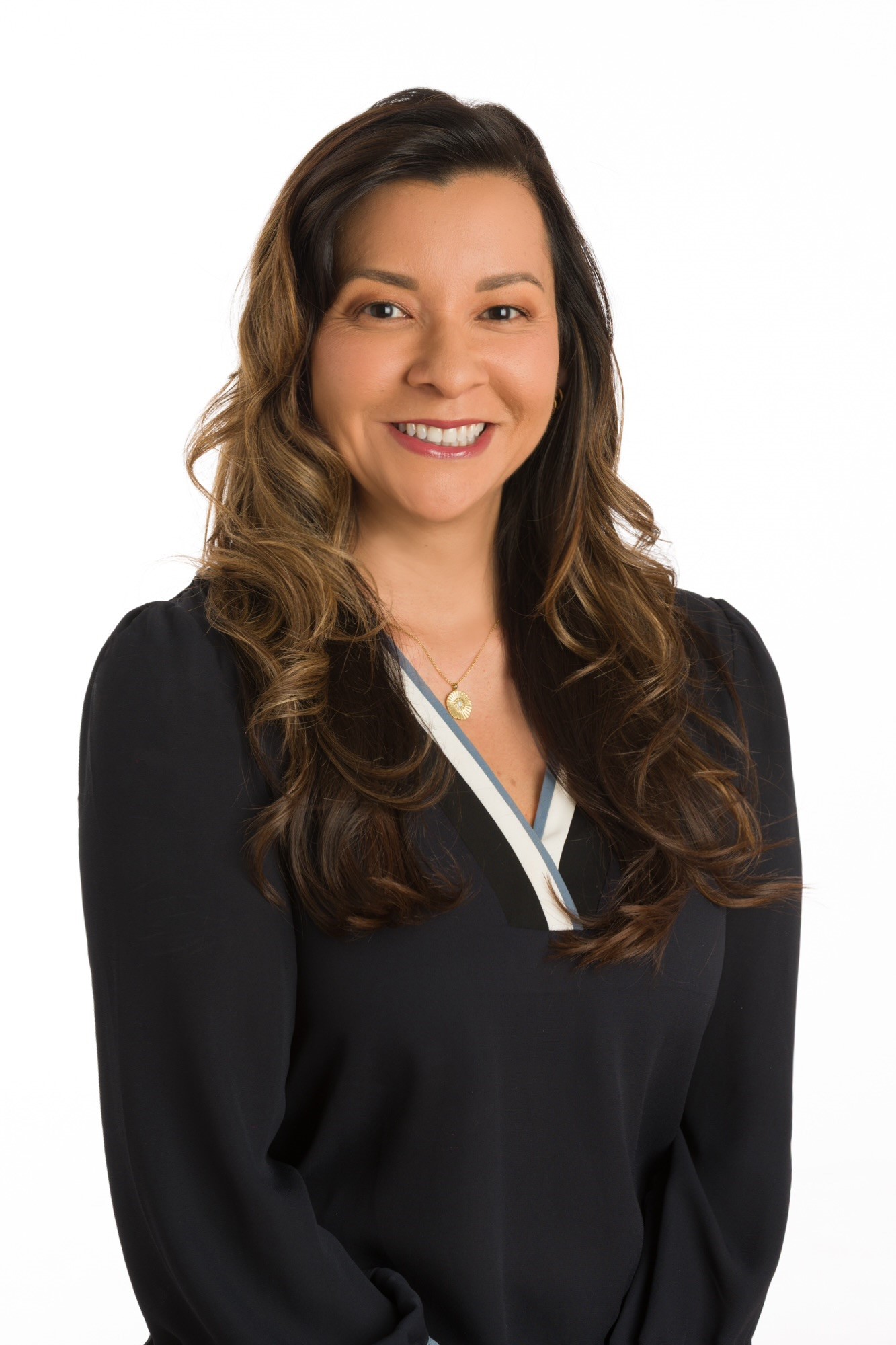 A woman with long wavy brown hair, wearing a dark blouse, stands in front of a plain white background, smiling at the camera.