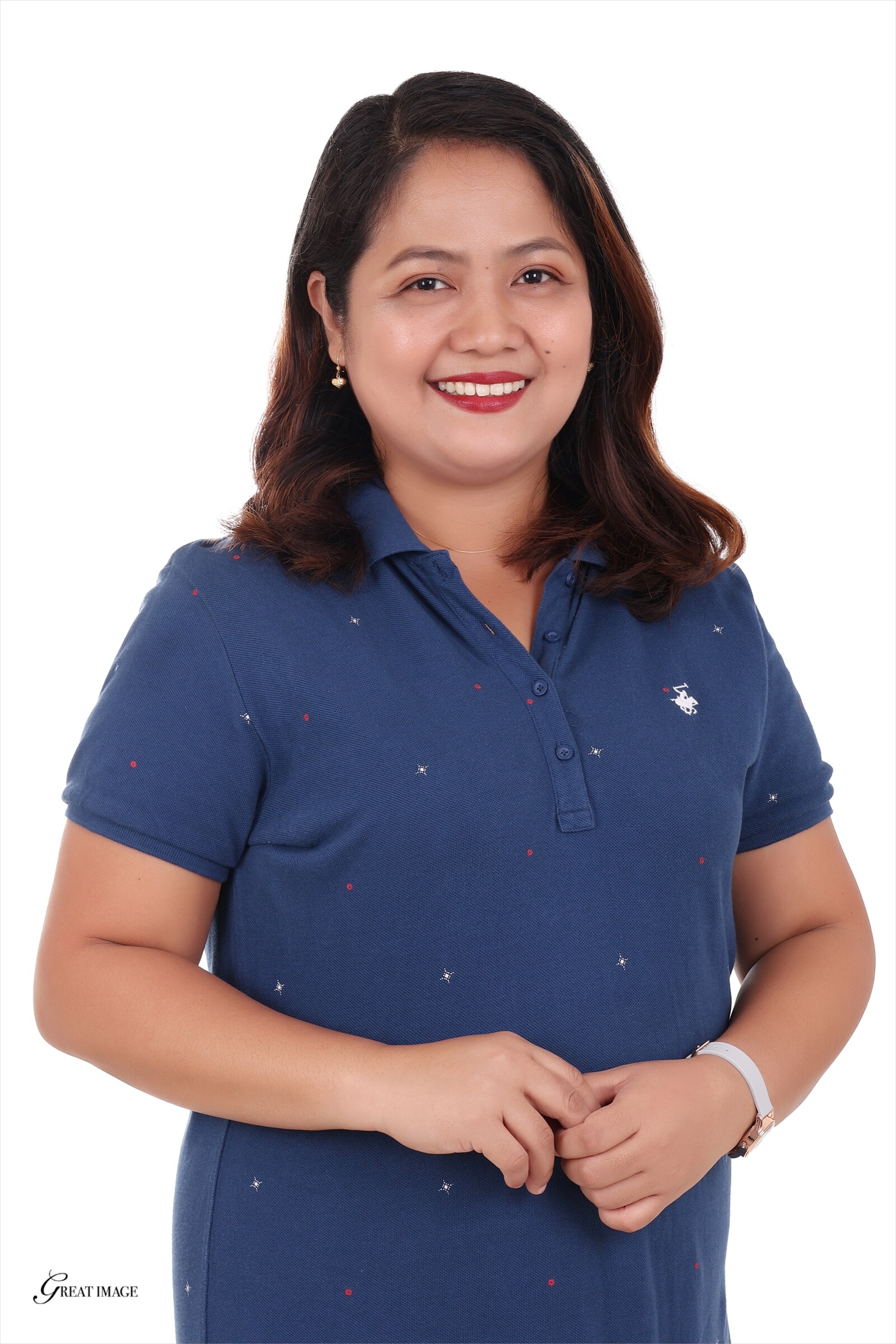 Melody Mayores, with shoulder-length hair, stands against a white background in a blue collared shirt, smiling warmly with her hands loosely clasped.