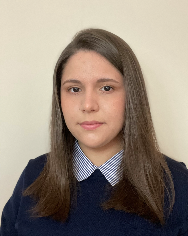 Person with long brown hair and a neutral expression, wearing a blue sweater over a striped shirt, standing against a plain light background.