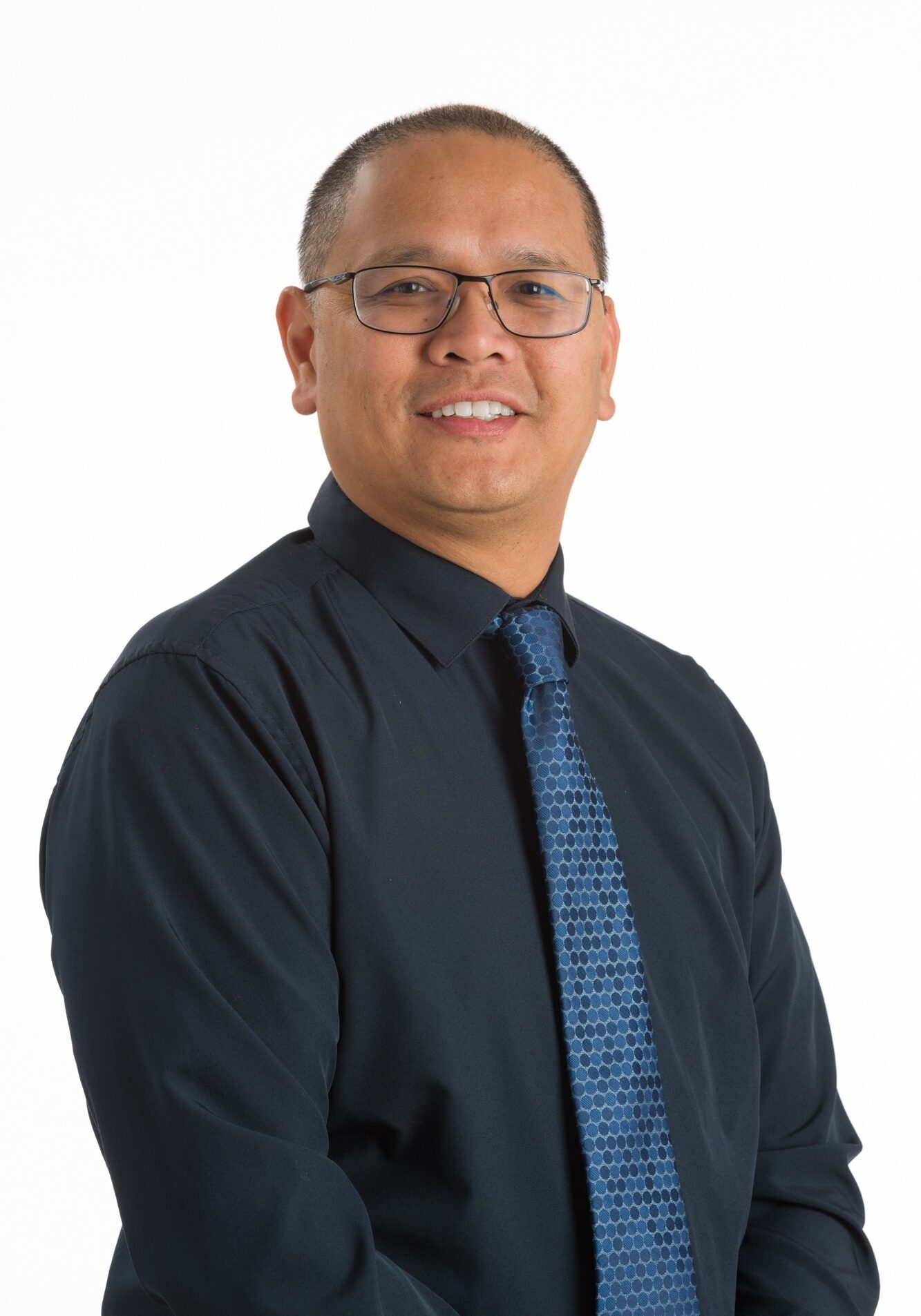 A man with short hair and glasses, wearing a dark shirt and patterned blue tie, poses against a plain white background.