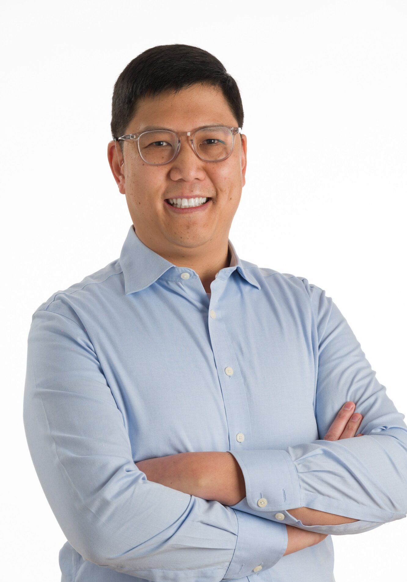 Christopher Chin, wearing glasses and a light blue button-up shirt, stands with arms crossed, smiling against a plain white background.