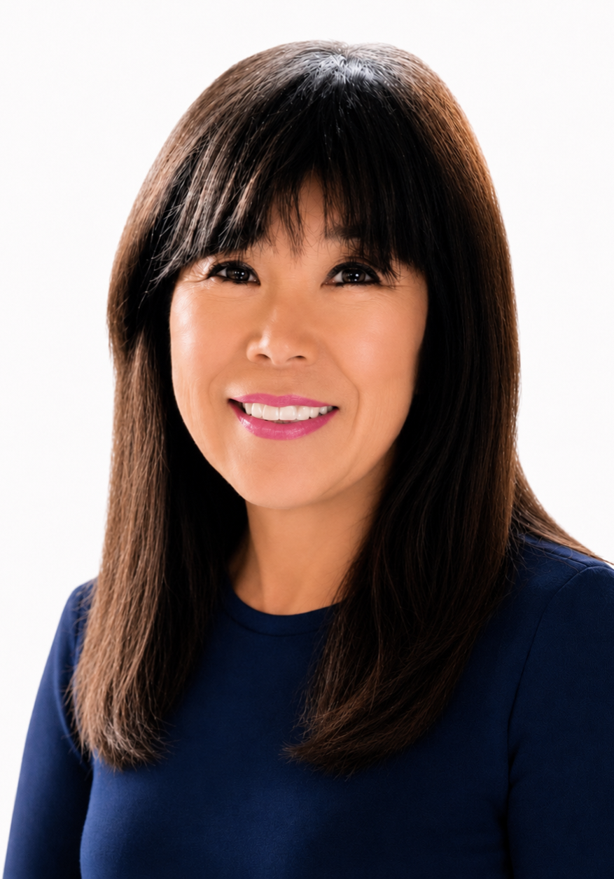 A woman with straight dark hair and bangs, wearing a navy blue top, smiles in front of a plain white background.