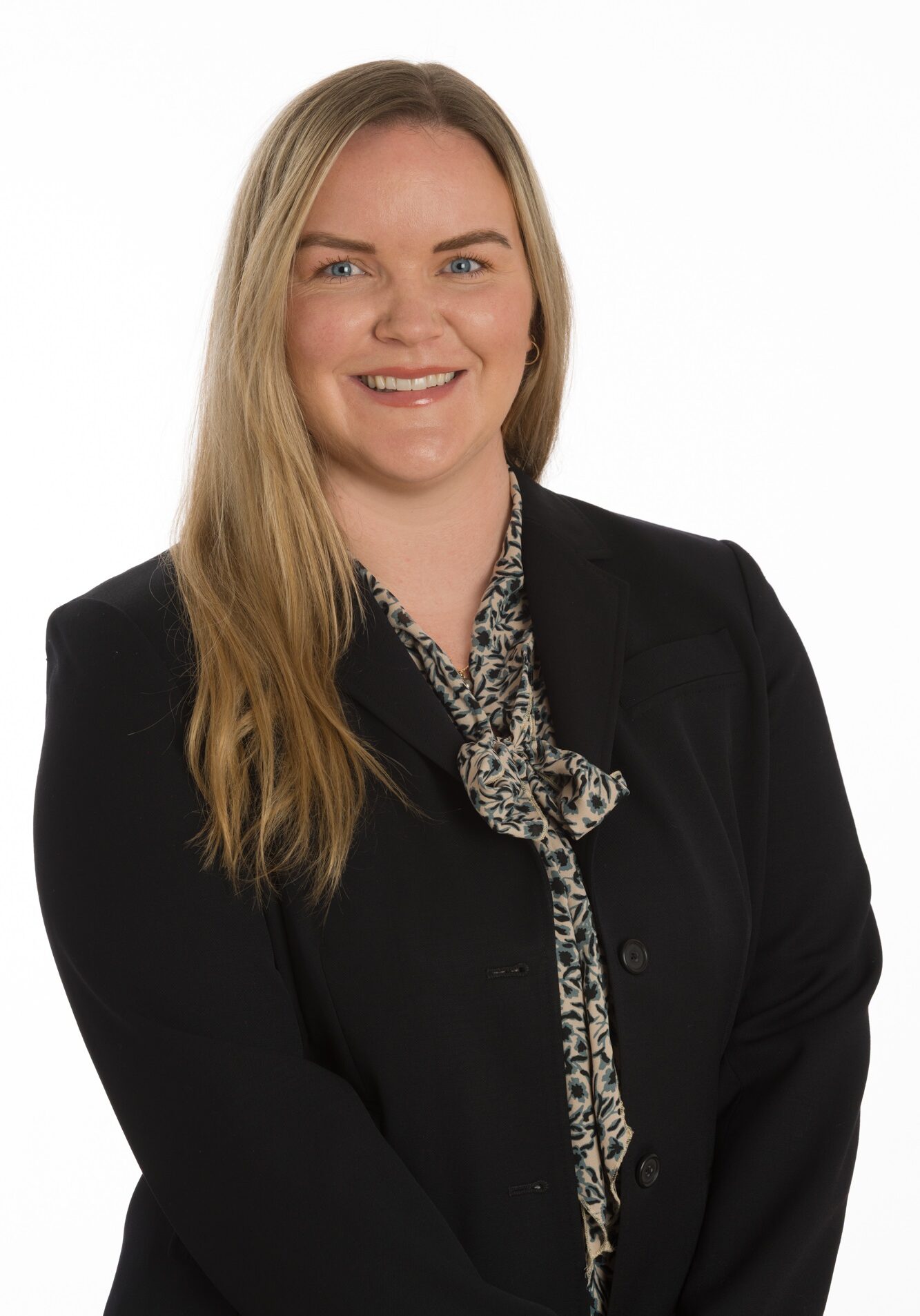 A woman with long blonde hair, wearing a black blazer and patterned blouse, smiles at the camera against a plain white background.