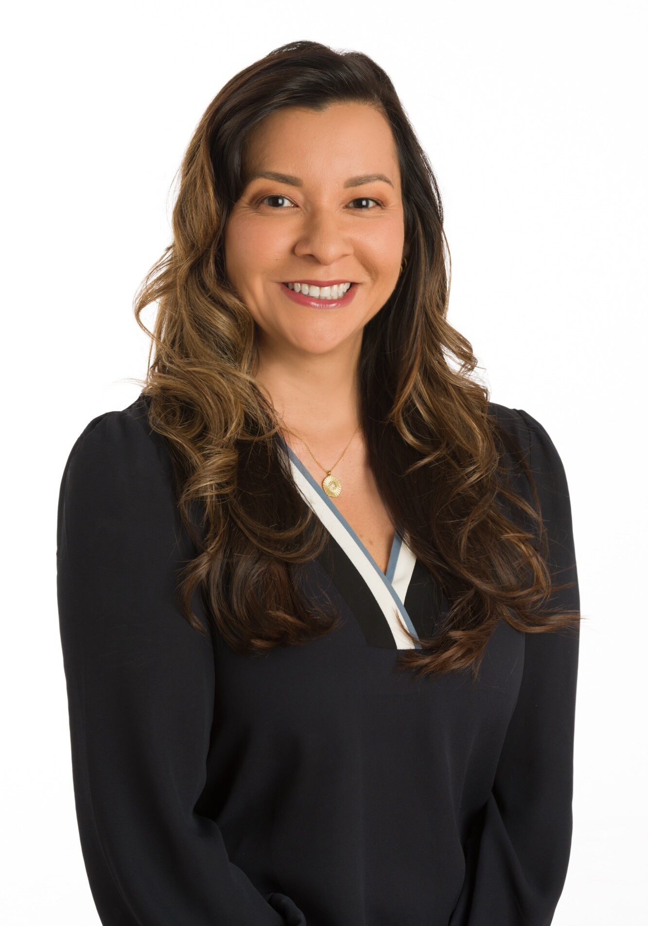 A woman with long wavy brown hair, wearing a dark blouse, stands in front of a plain white background, smiling at the camera.